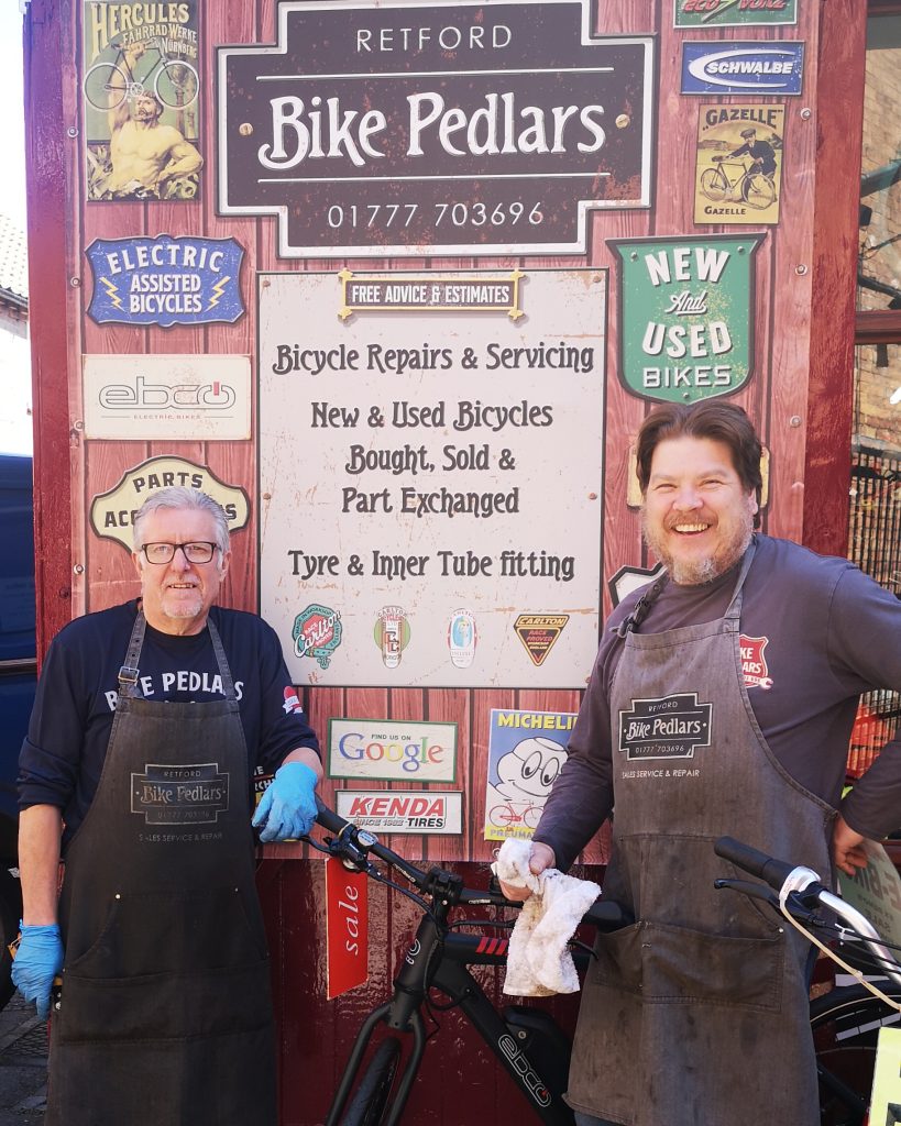 Two smiling mechanics in aprons stand in front of a colorful wall outside "Bike Pedlars" in Retford, which advertises bicycle repairs, sales, and servicing. The backdrop features vintage-style signs, tire brand stickers like Michelin and Kenda, and services listed such as “New & Used Bicycles Bought, Sold & Part Exchanged” and “Tyre & Inner Tube fitting.”